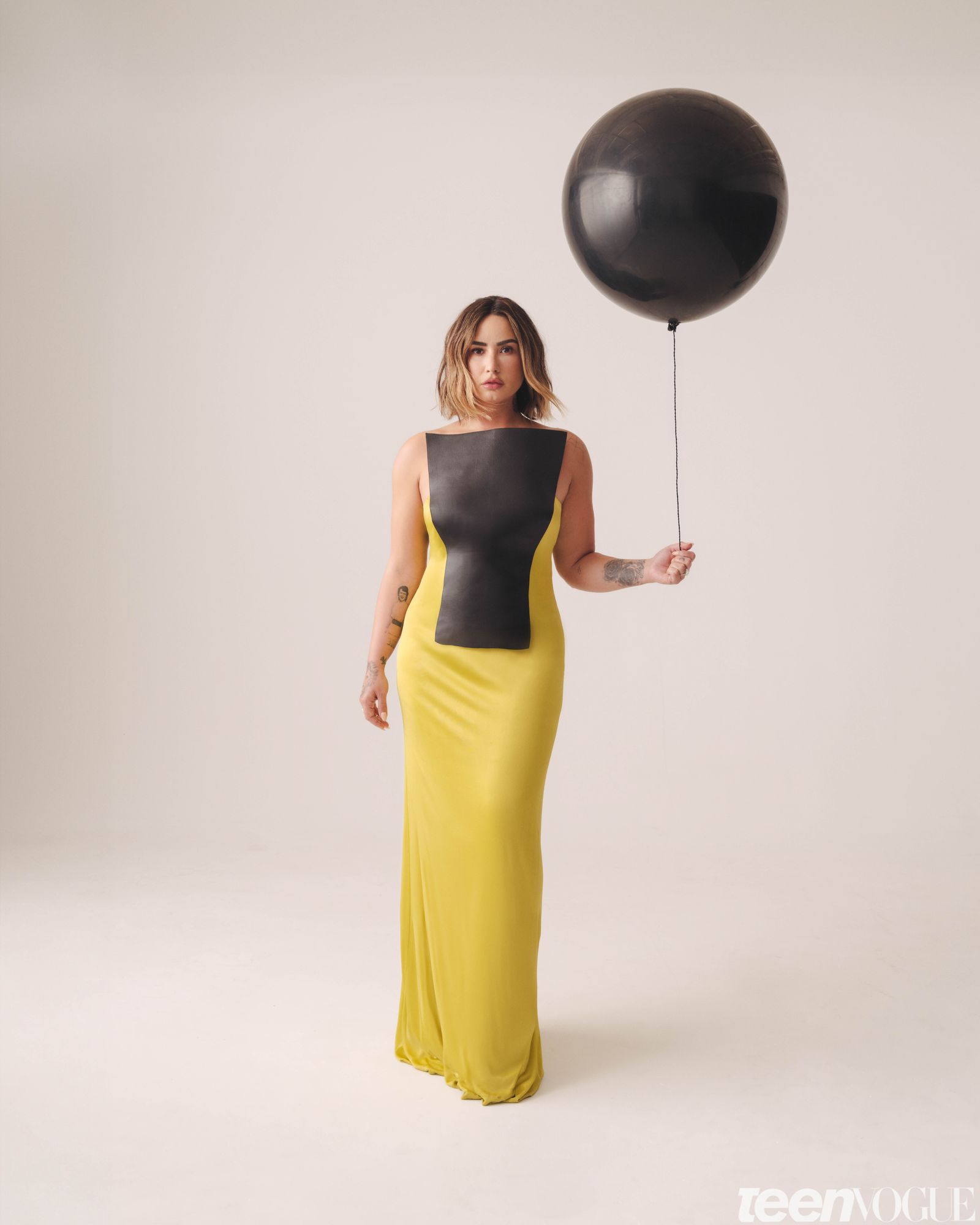 Demi gazing directly towards viewer hair softly blowing in the wind wearing a black and yellow bodycon floorlength gown...