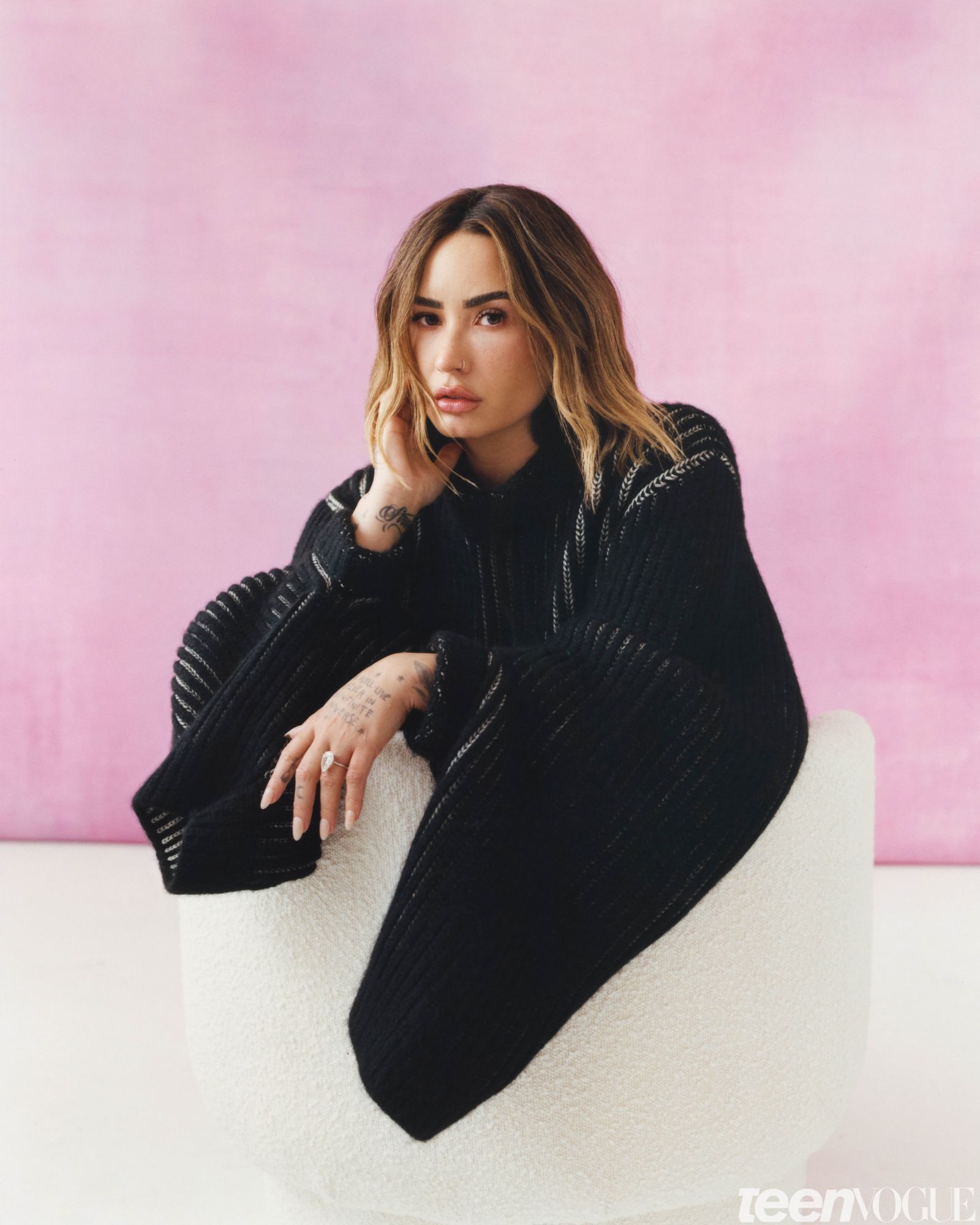 Demi's hand touching face and other resting on chair looking directly towards viewer wearing an oversized black knit...
