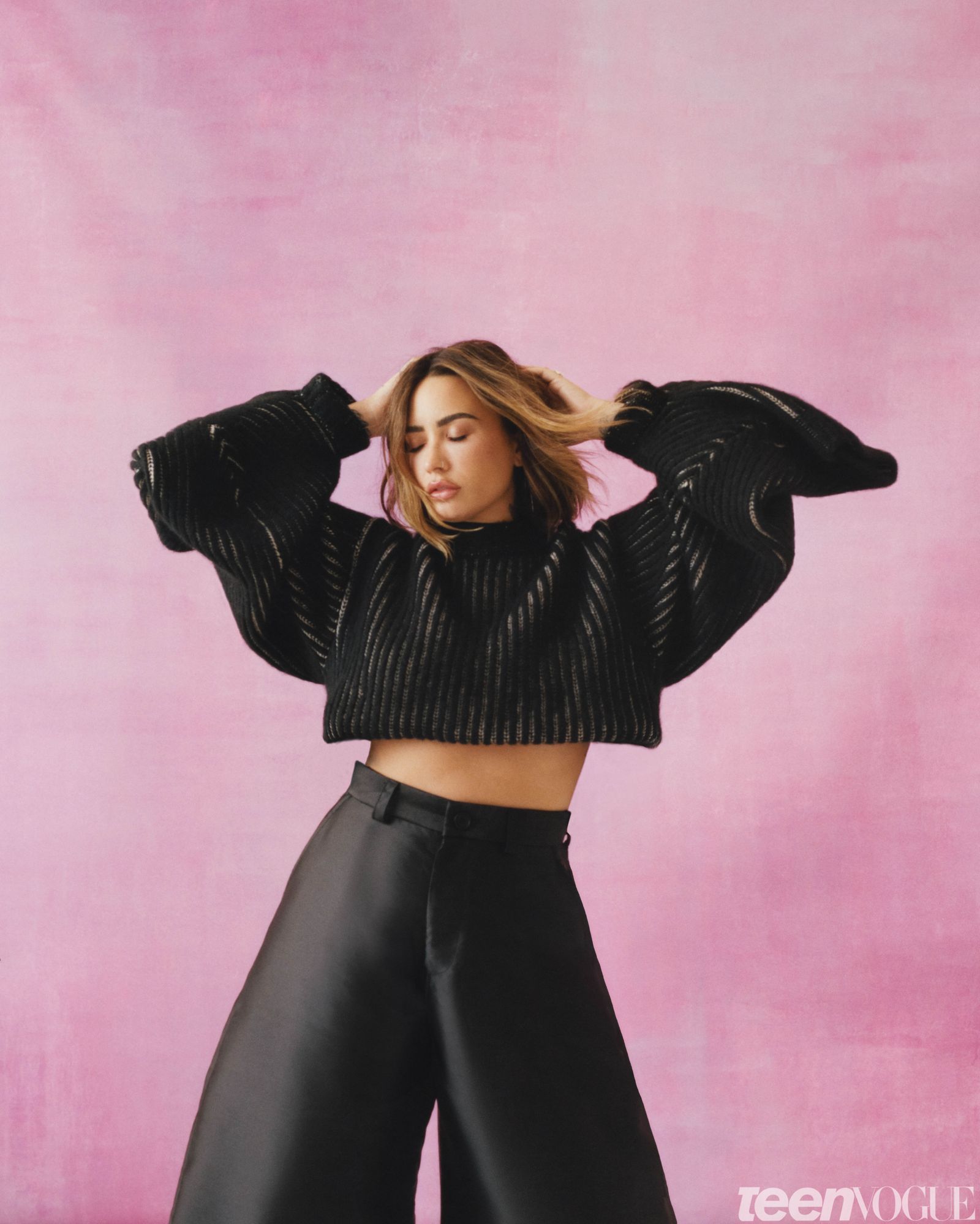 Demi wearing a black knit sweater and black dress pants hair and body swaying against a pink backdrop.