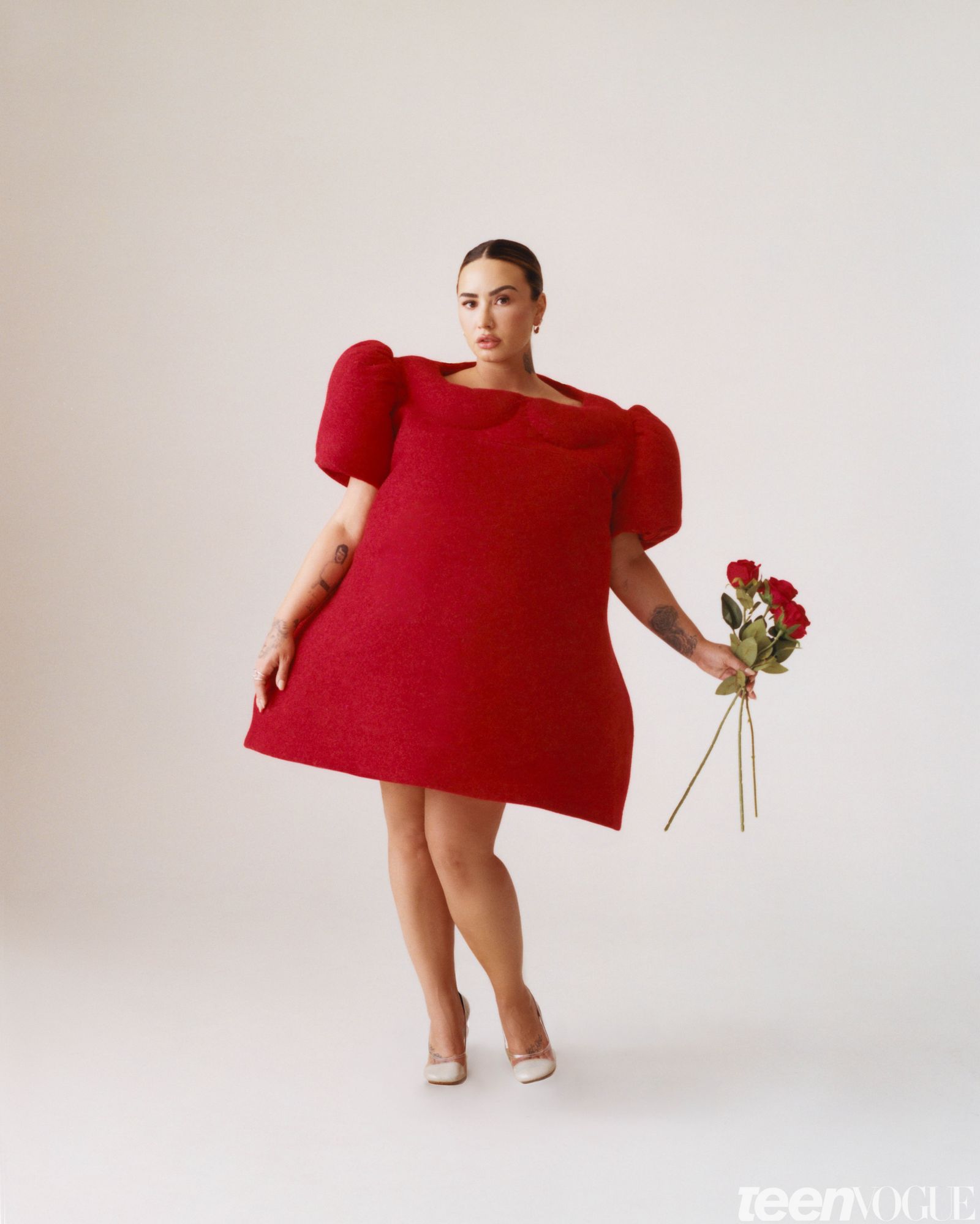 Demi photographed in a playful structured doll like red dress holding three red roses.
