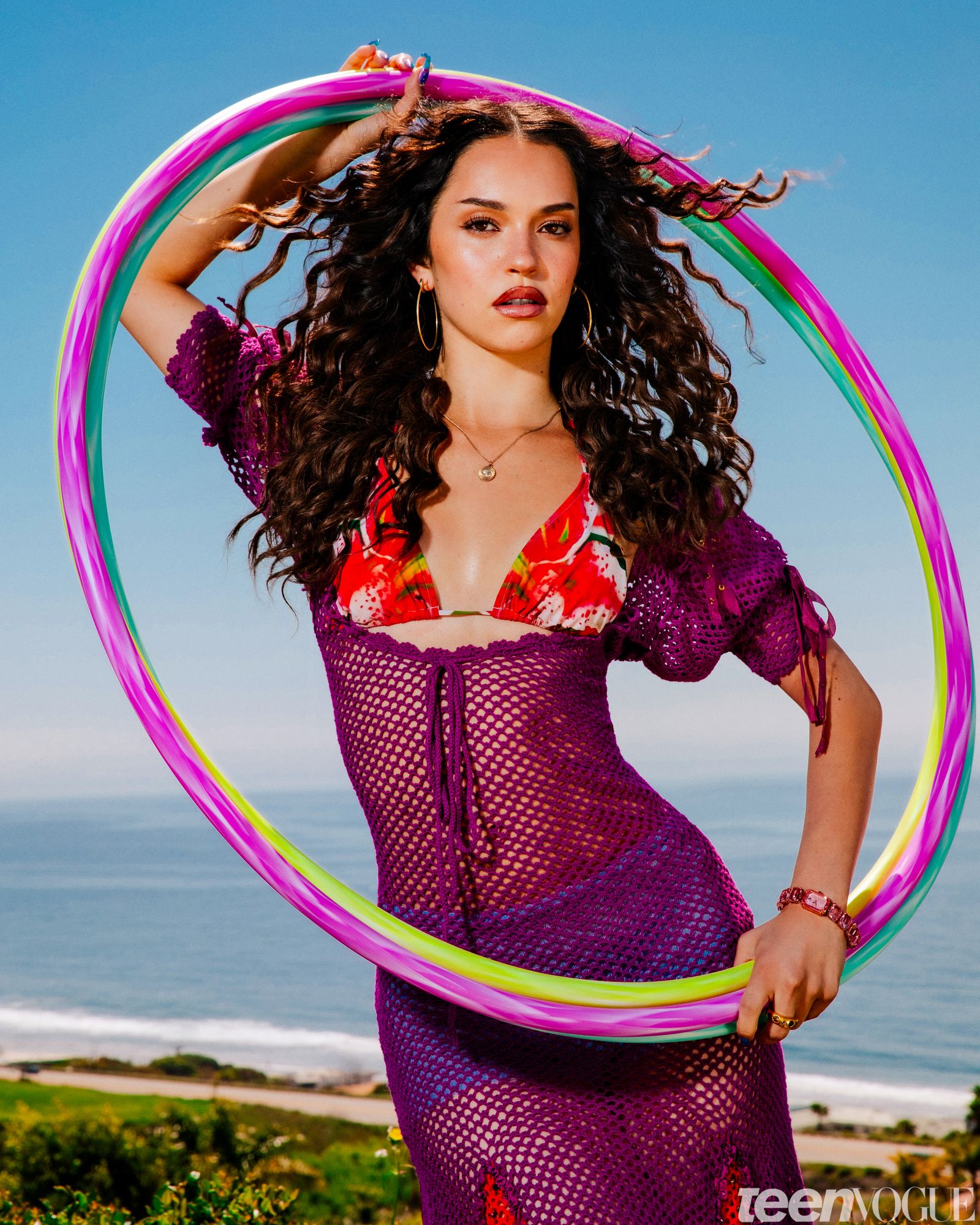 Dani of KATSEYE photographed outside holding hula hoops framing her face. She wears a woven purple dress.