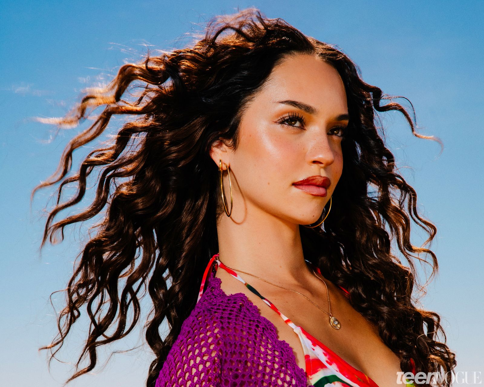 Dani photographed against the blue sky in purple woven dress and red tropical print bikini top curls flowing in the wind.