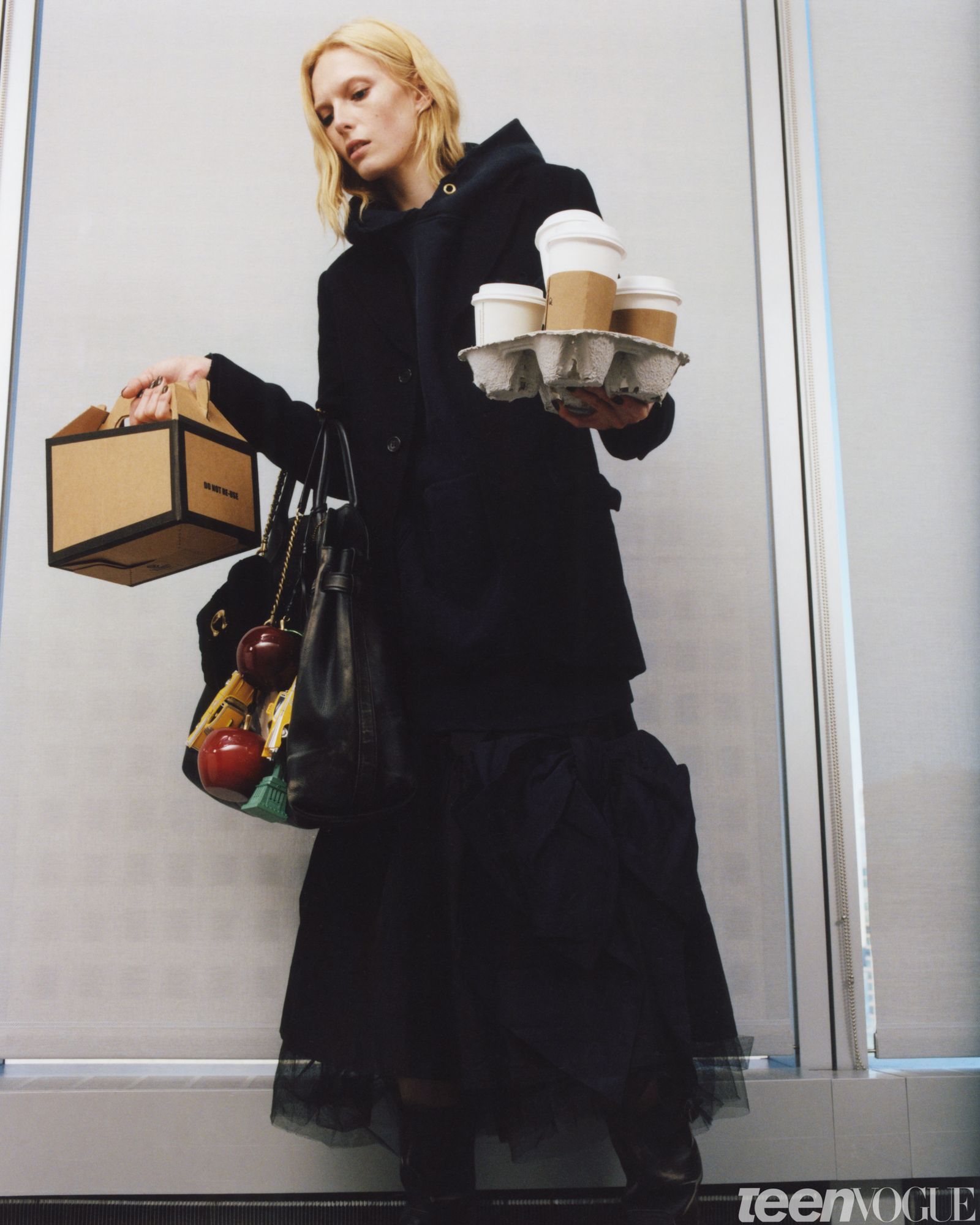 Alex as the Production Assistant. She looks away from viewer holding a tray of disposable coffee cups in one hand and a...