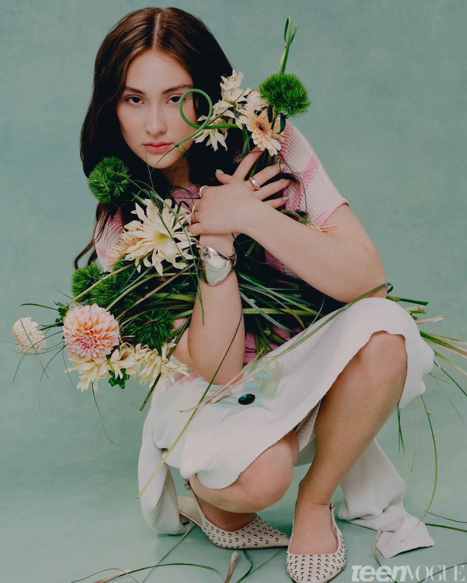 Lola Tung photographed crouching down directly facing viewer holding a bouquet of pastel flowers with both hands while...