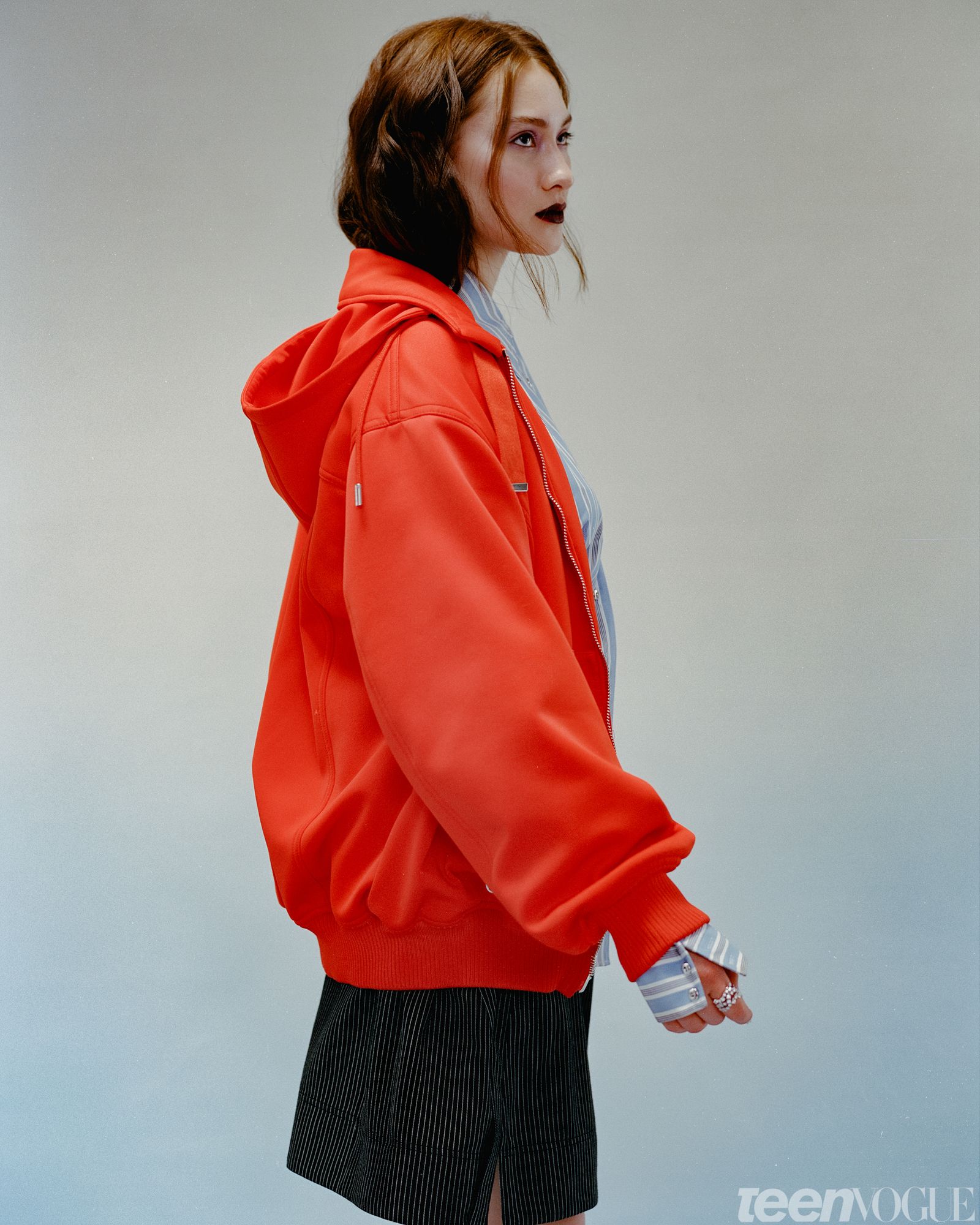 Lola Tung photographed standing against a teal backdrop looking away from viewer wearing a bright orangered hoodie. She...