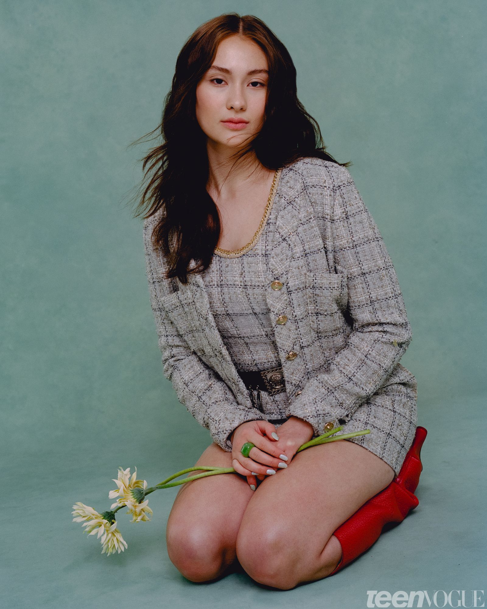 Portrait of Lola Tung kneeling looking at viewer gently holding two pastel yellow flowers against a mint green backdrop....
