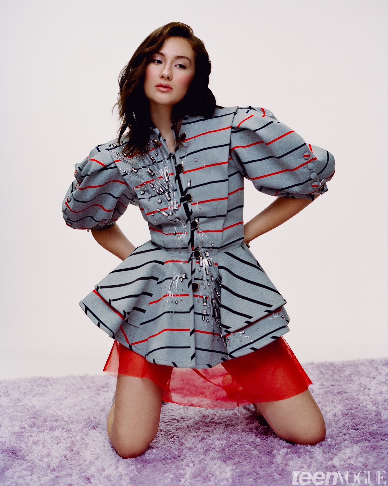 Lola Tung photographed against a white backdrop kneeling on a pastel purple rug with her hands on her hips. She looks...