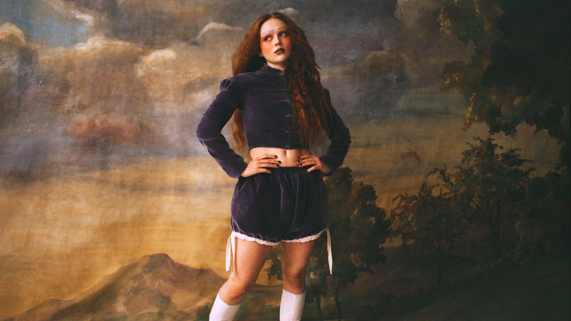 Sadie Sink with her hands on her hips wearing a purple suede twopiece set  and white boots.
