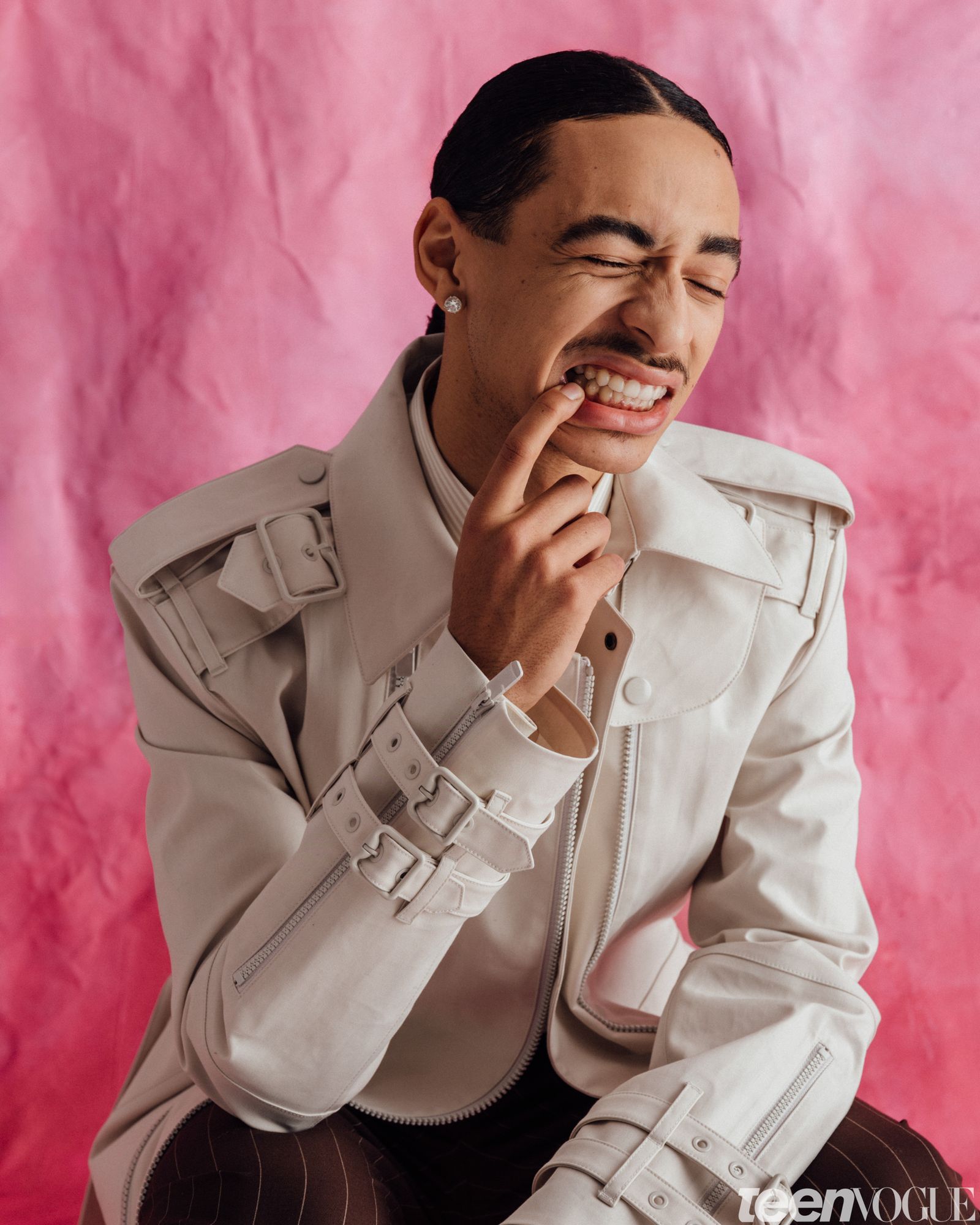 Julez pulling a grill pose and grinning while kneeling in front of a pink backdrop wearing a structured jacket.