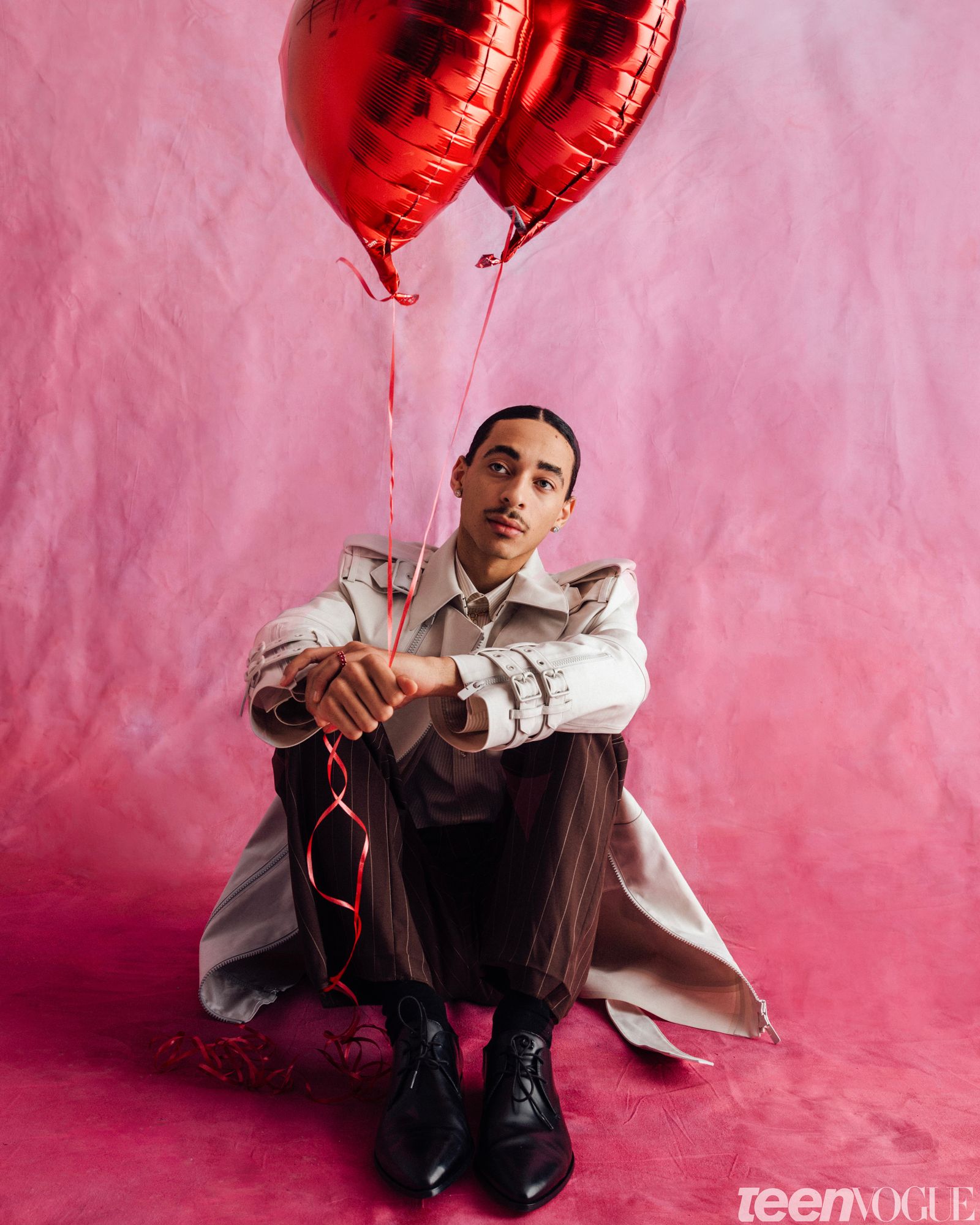 Julez sitting on a pink backdrop wearing a structured jacket and striped brown trousers while holding two mylar red...