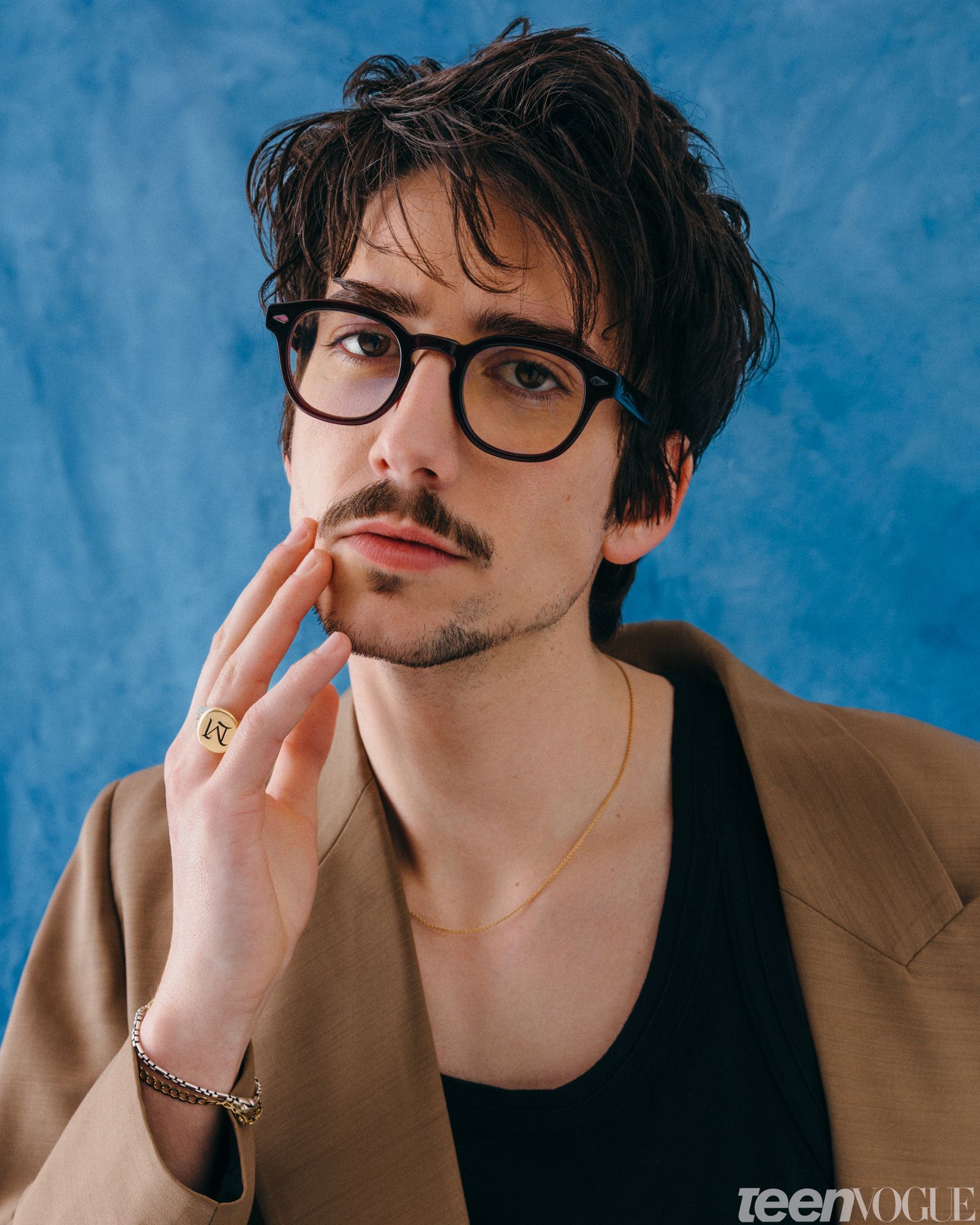 Milo glasses on wearing a tan suit and black tank top touching the side of his face gazing directly at viewer.