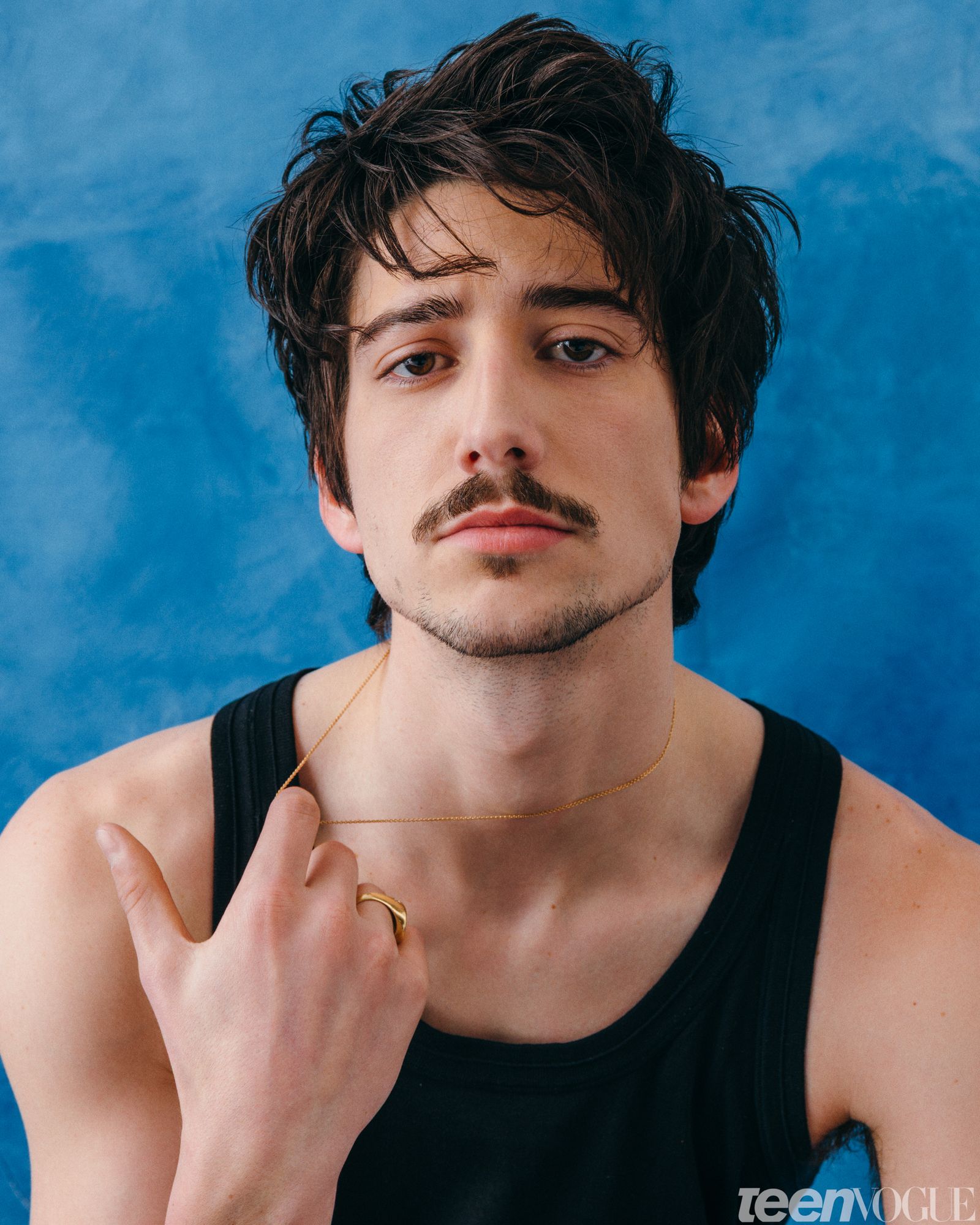 Milo in a black tank top holding the gold chain around his neck gazing directly towards viewer.