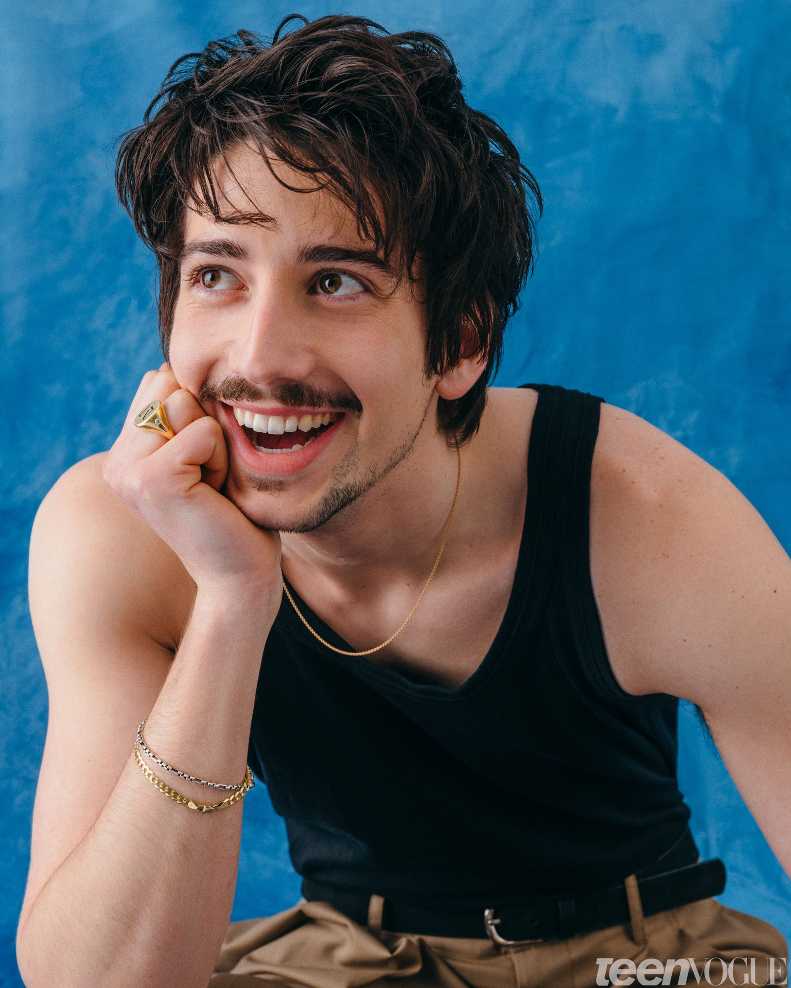 Milo smiling from ear to ear while looking away from viewer with arm resting on knee with in a clutched fist against his...