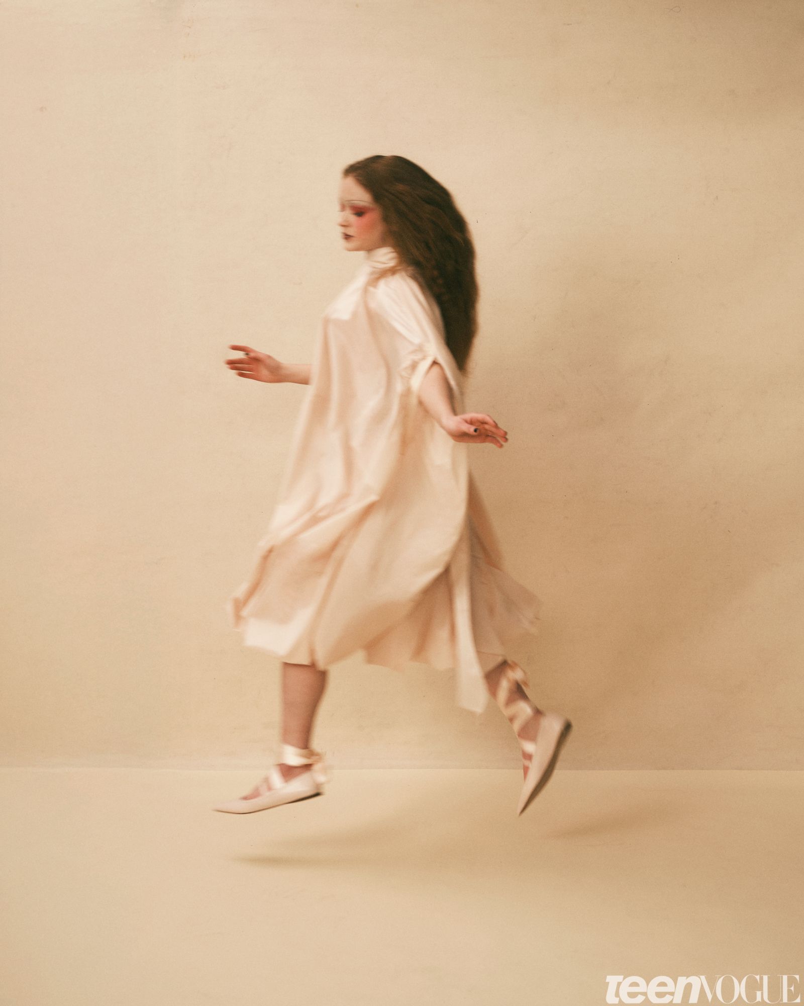 Sadie Sink leaping with her hands out by either side against a beige background wearing a flowy dress and ballet flats...