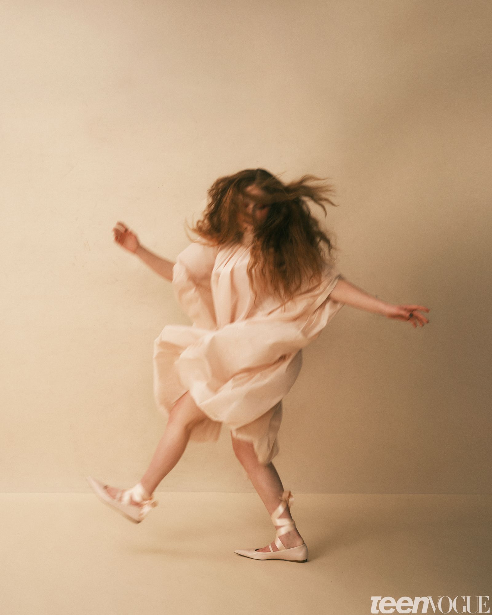 Sadie Sink in motion with her hands out by either side hair covering her face against a beige background wearing a flowy...