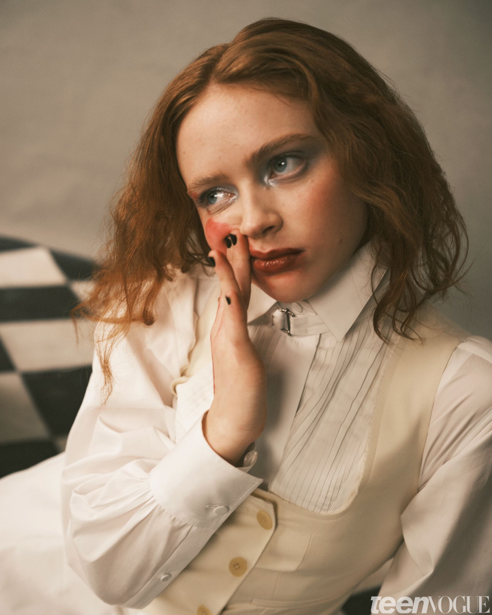 Sadie smudges her lipstick with her right hand. She is sitting on a blackandwhite checkered floor and wearing a collared...