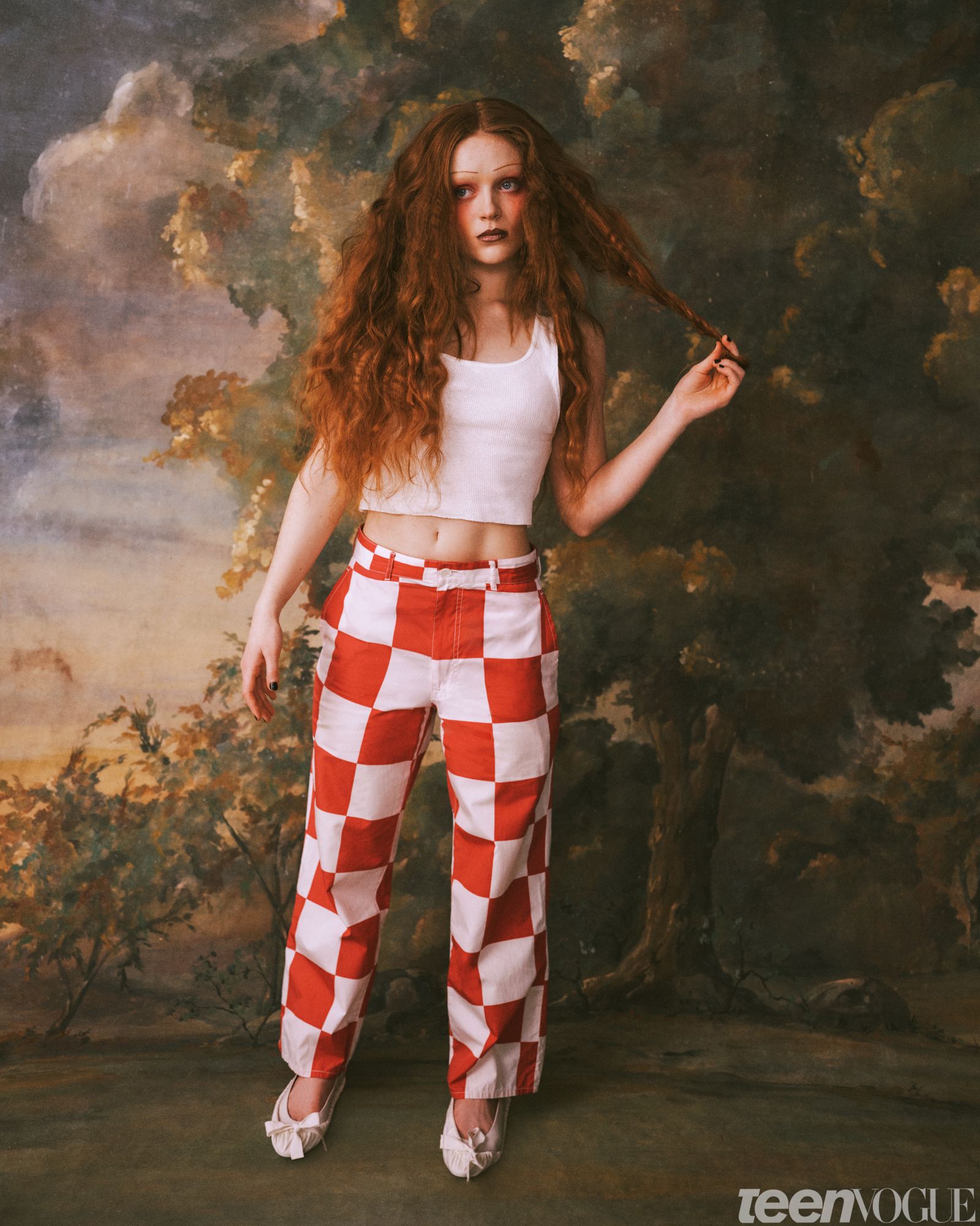 Sadie playing with her hair while in a cropped white tank top and pinkandwhite checkered pants against a painted...