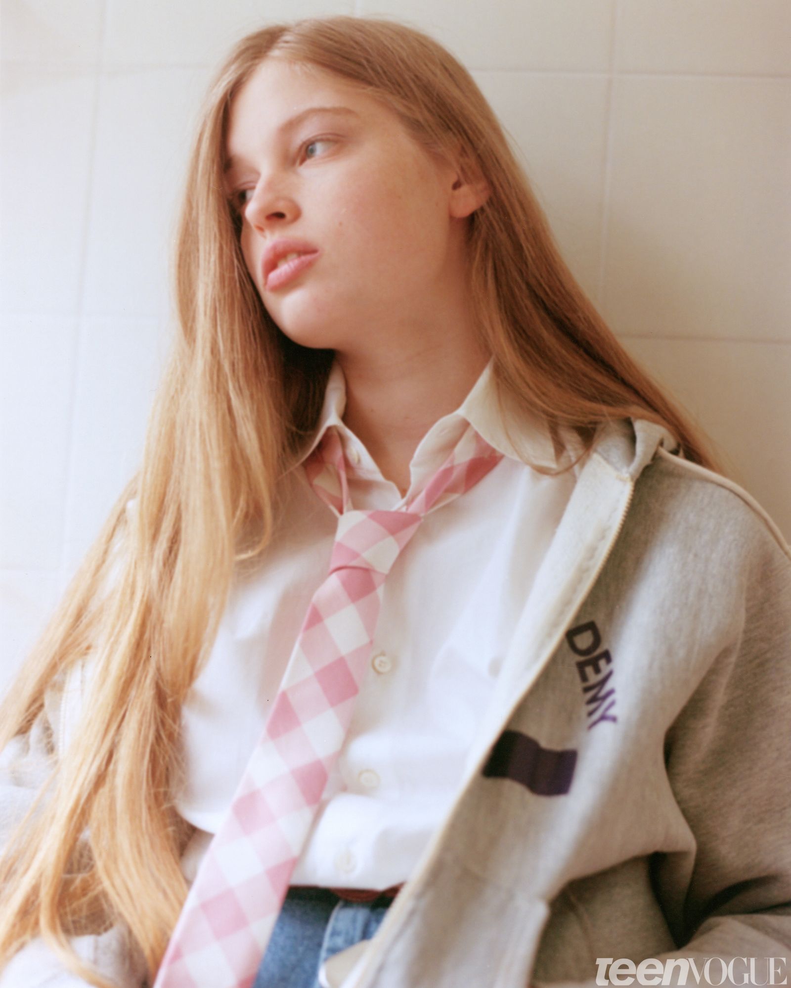 Closeup portrait of Vivian looking away from viewer against a cream tiled wall. She wears a pink and white gingham tie...