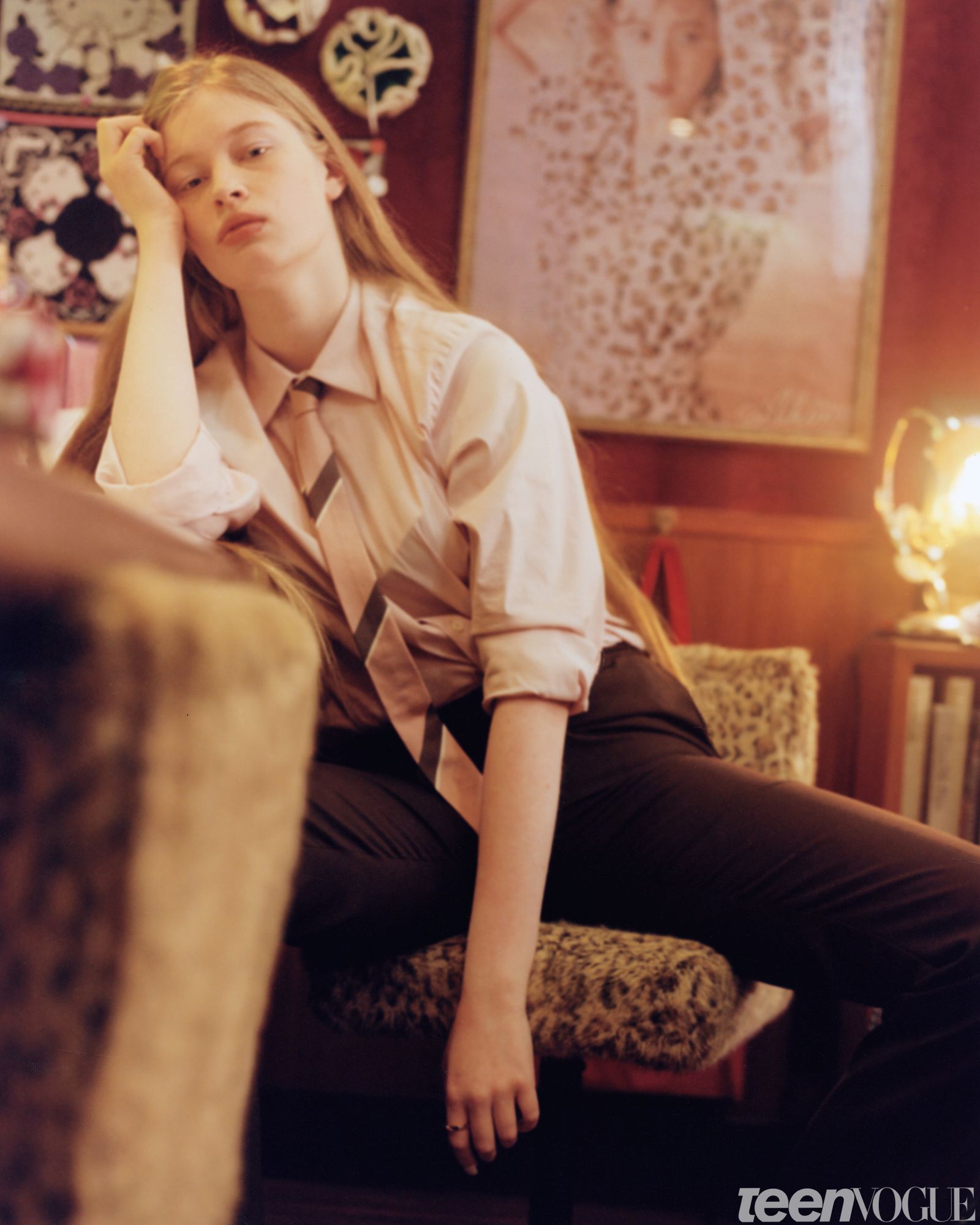 Vivian looks away from viewer while sitting hunched over on a leopard print chair with her hand resting on her face. She...