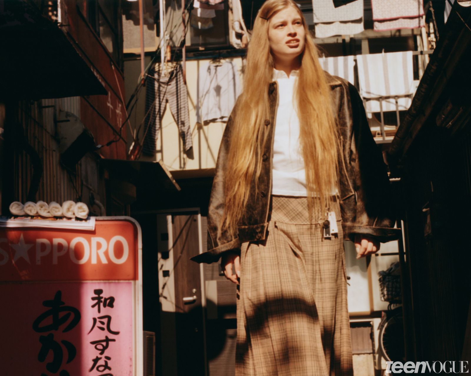 Vivian walking through a Tokyo alley while wearing an oversized biker jacket white shirt plaid skirt and black loafers.