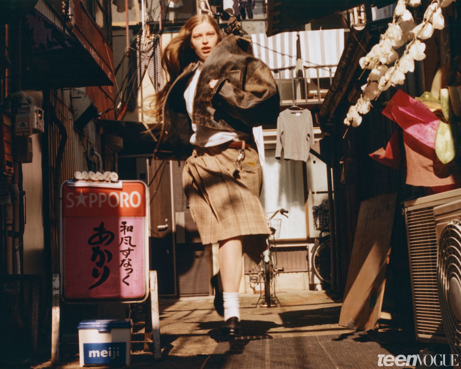 Vivian running through a Tokyo alley while wearing an oversized biker jacket white shirt plaid skirt and black loafers.