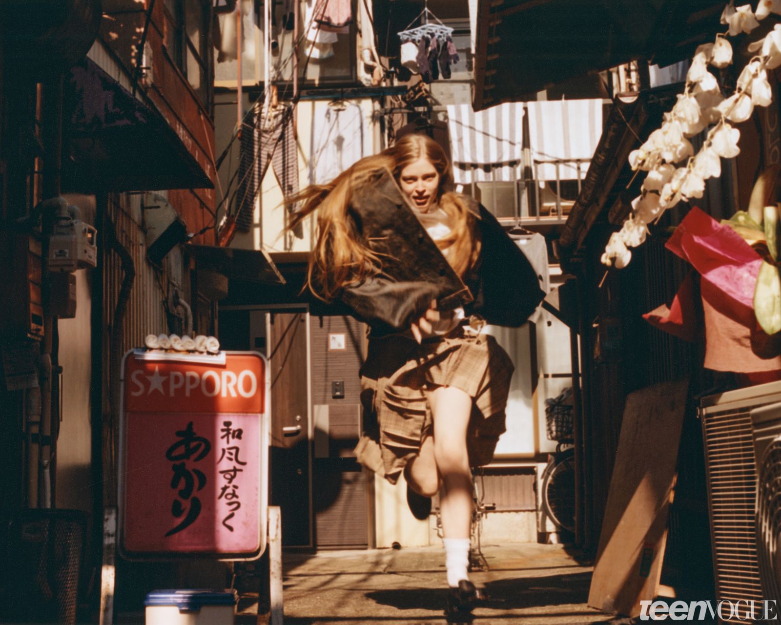 Vivian running through a Tokyo alley while wearing an oversized biker jacket white shirt plaid skirt and black loafers.