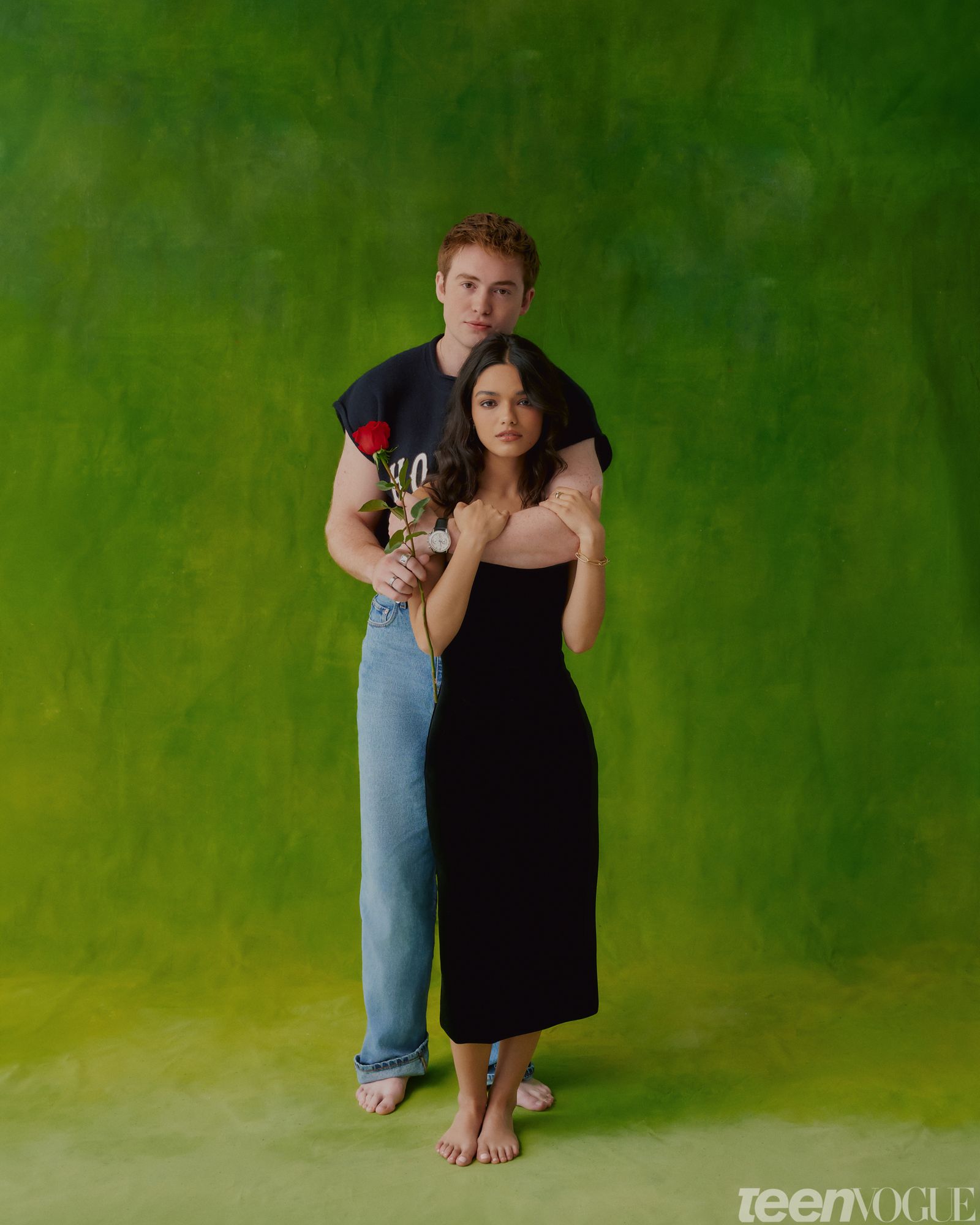 Kit Connor and Rachel Zegler standing together against a green backdrop. Kit's arm around Rachel holding a red rose.