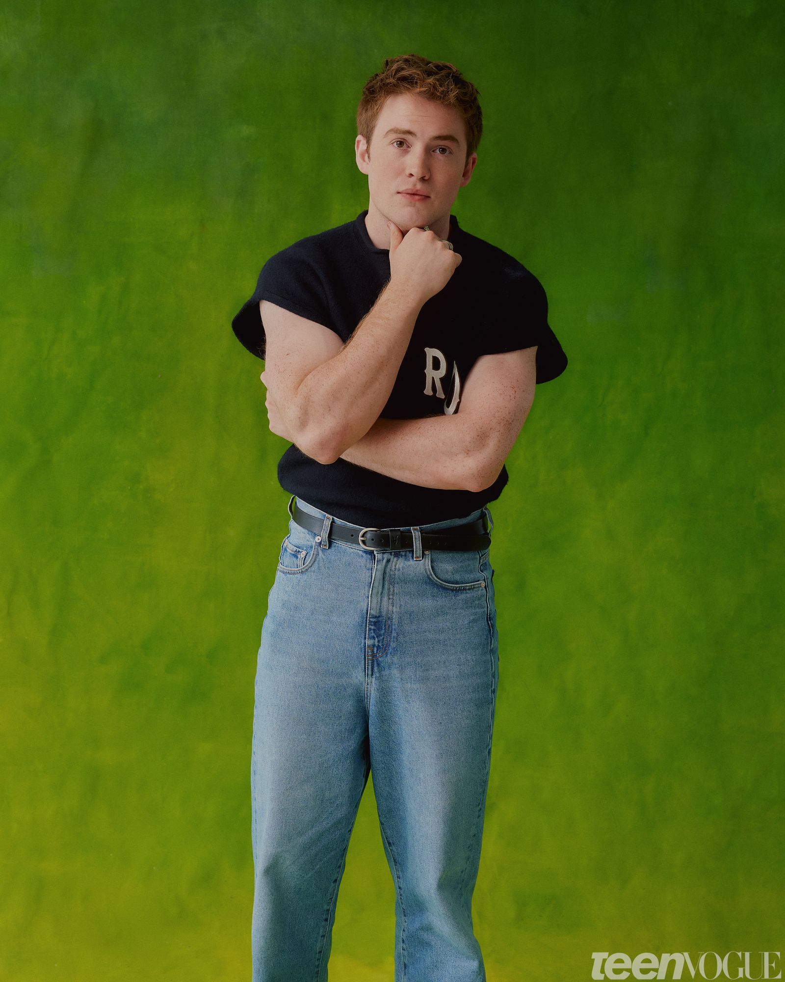Kit Connor standing against a green backdrop wearing a navy sweatshirt and jeans.